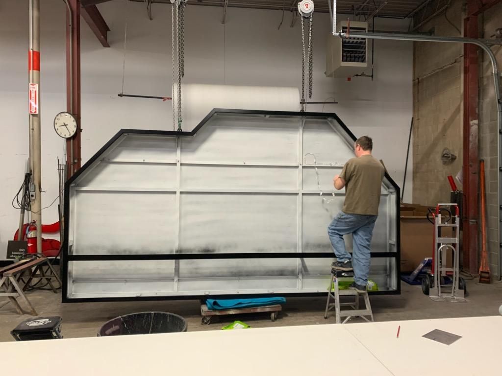 Man on step stool working on a large white rectangular frame with black trim; industrial setting.