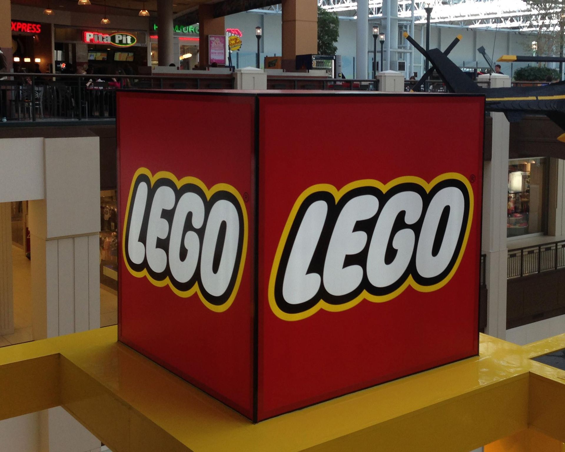 Large red LEGO cube with white text and yellow border, in a mall setting.