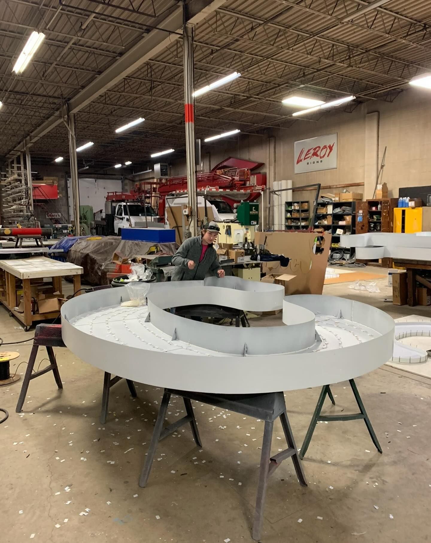 Man in workshop near a large, circular, light-colored sign under construction.