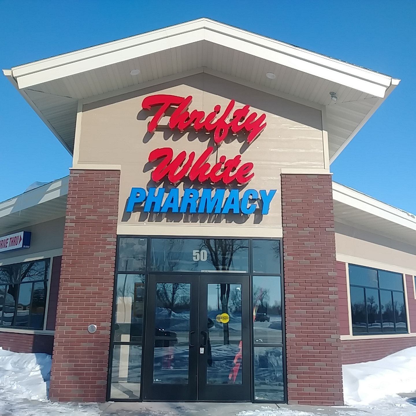Thrifty White Pharmacy storefront with red and blue sign, brick and glass facade, snow on the ground.