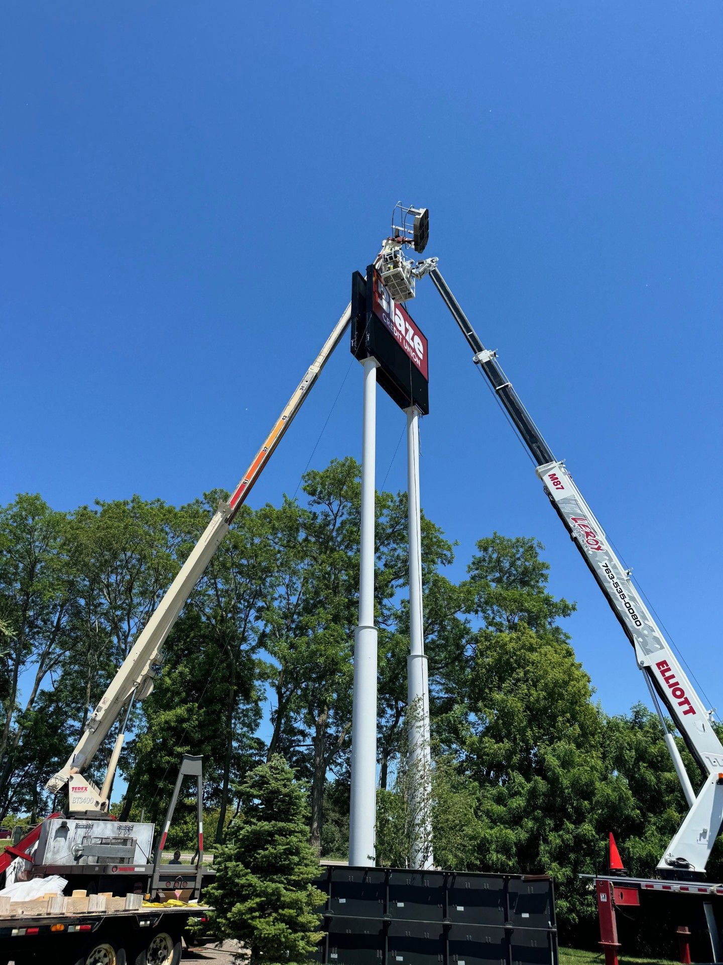 A crane lowering a sign with a logo onto a pole; a worker on a lift is guiding it. Cloudy sky.