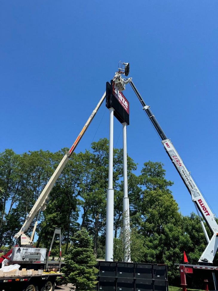 A crane lowering a sign with a logo onto a pole; a worker on a lift is guiding it. Cloudy sky.