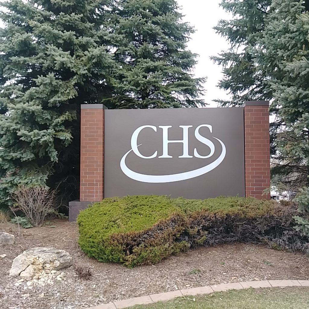Sign for CHS with white letters and a swoosh on a brown background, flanked by brick pillars and evergreen trees.
