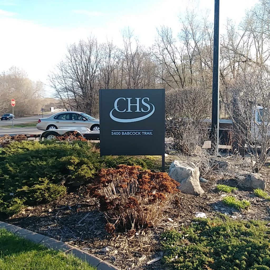 CHS sign, black with white logo, in front of bushes and trees; cars on road in background.