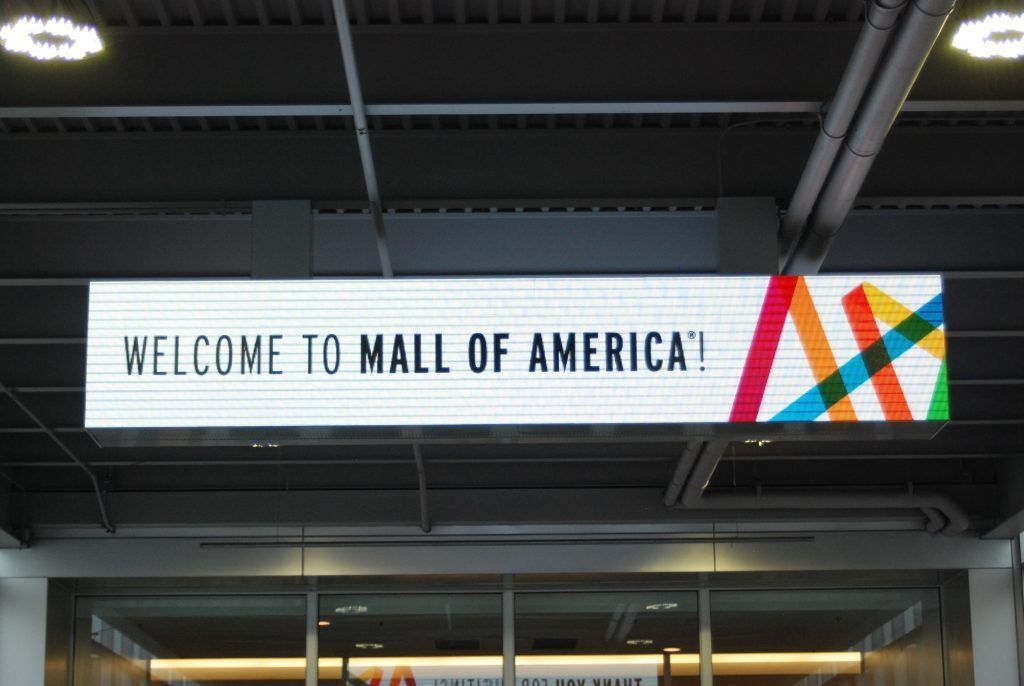 Sign reading Welcome to Mall of America! with colorful logo, suspended from ceiling.