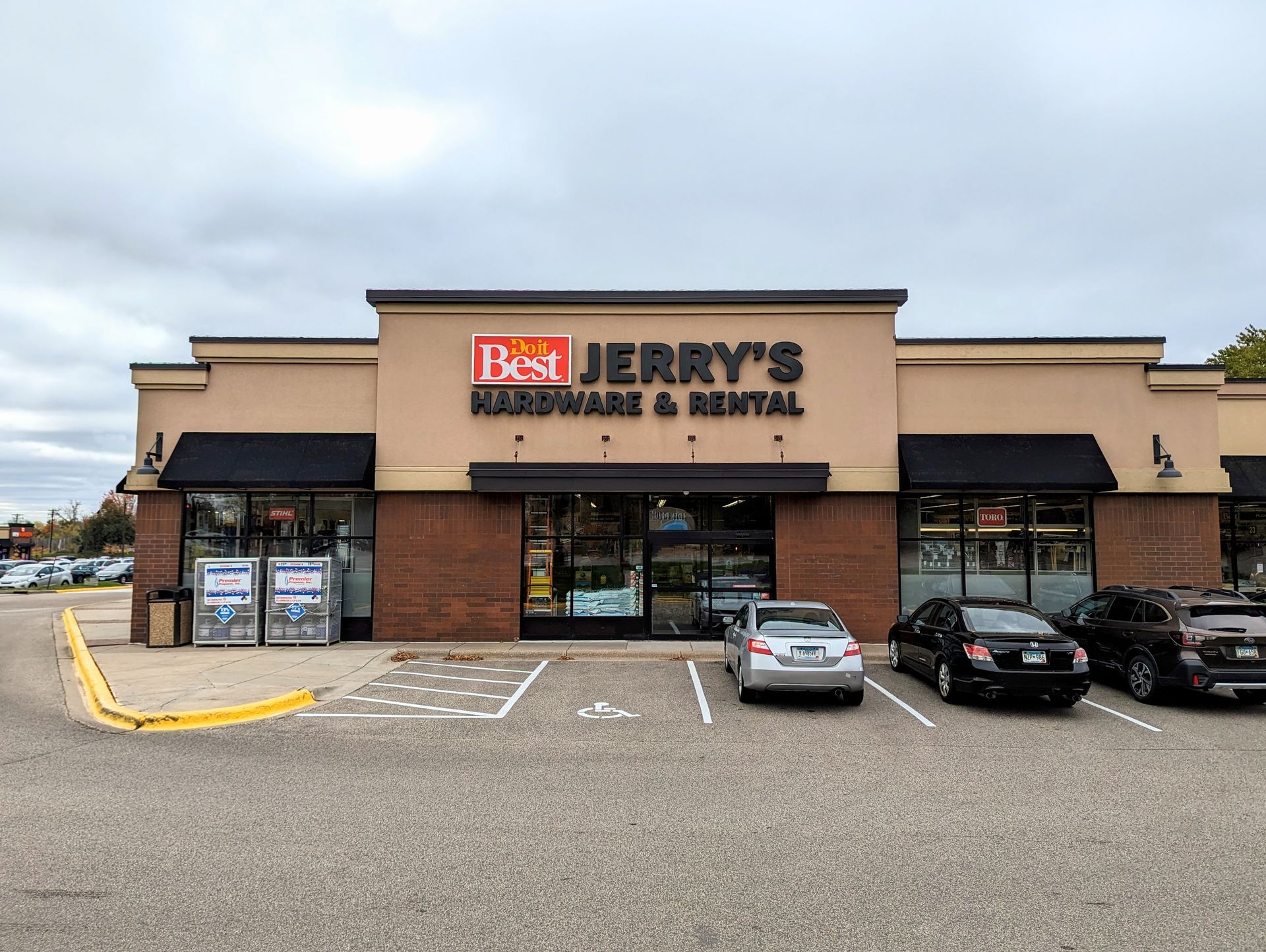 Best Jerry's Hardware & Rental store with tan facade, black awnings, and parked cars in front. Cloudy sky.