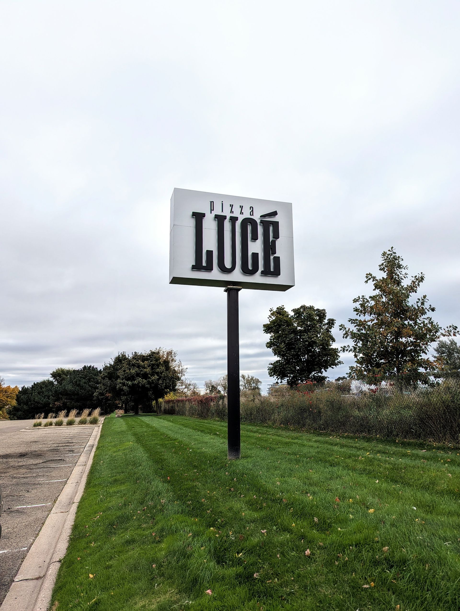 Sign for De Luce restaurant, black letters on white background. Set on green grass under overcast sky.