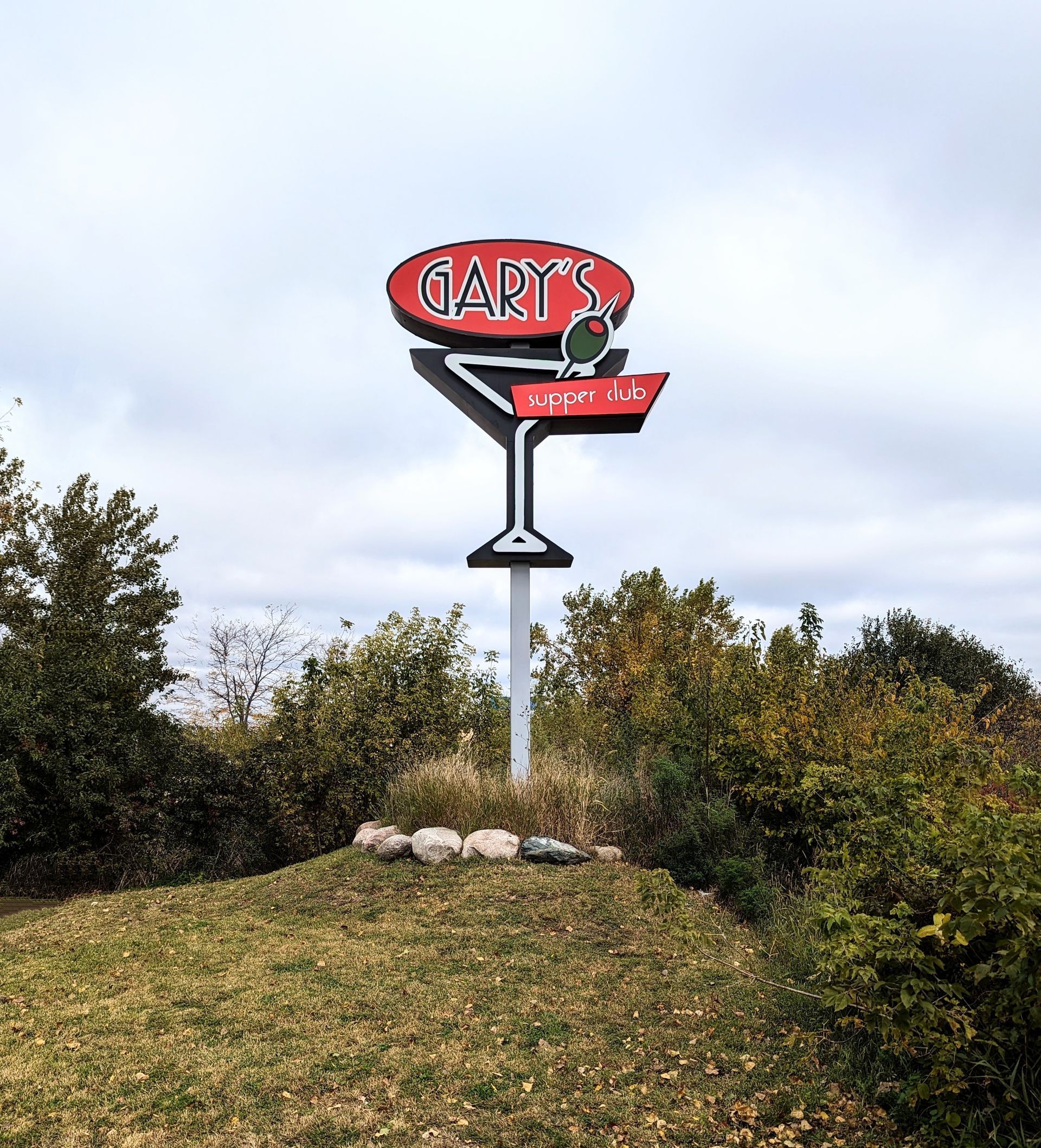 Gary's Bar sign, red and white, martini glass shape, on a hill with foliage.
