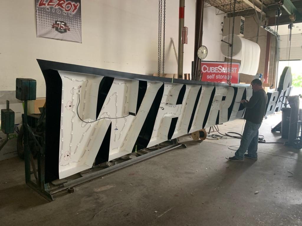 Man works on a large WHIRL sign with black trim and white letters inside a workshop.