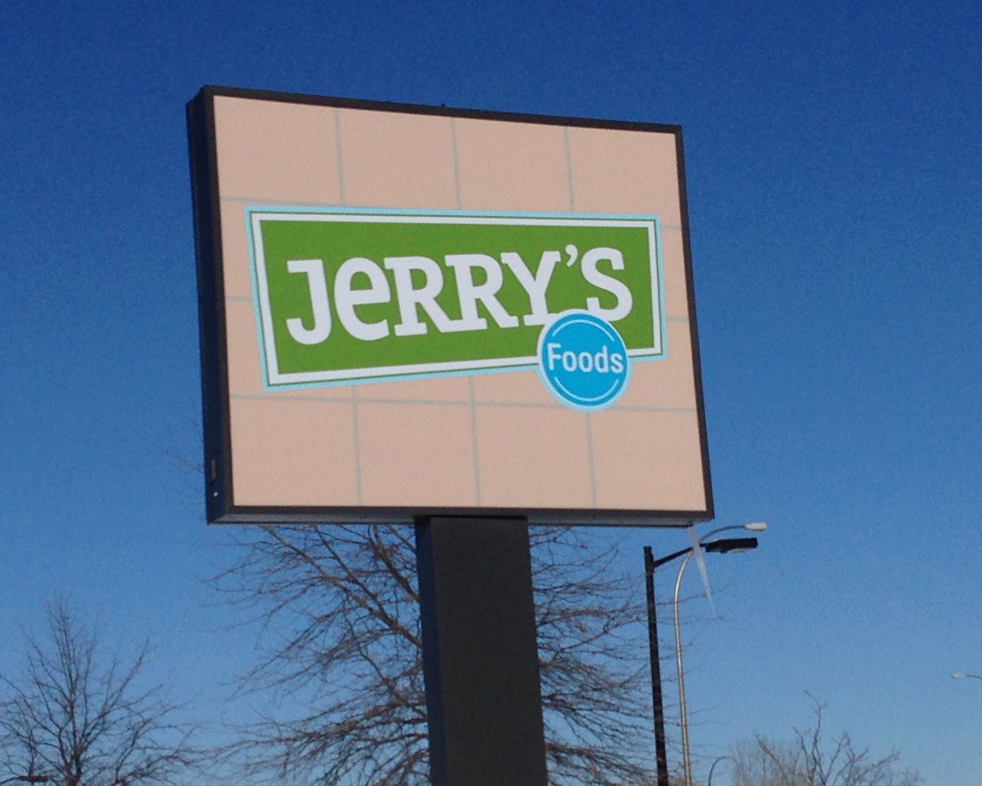 Jerry's Foods store sign, green text on a white background, with a light pink background, against a blue sky.