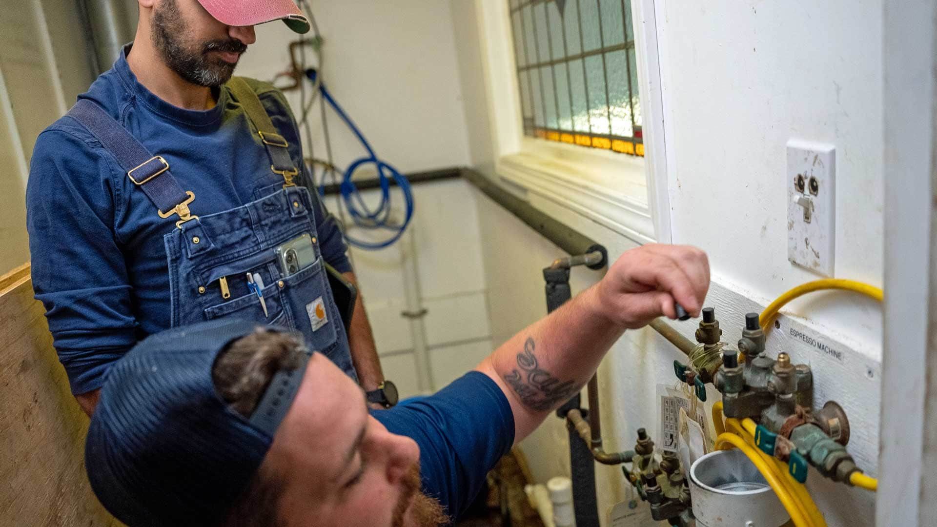 Plumber repairing a burst pipe, water spraying, wearing gloves and a blue beanie.