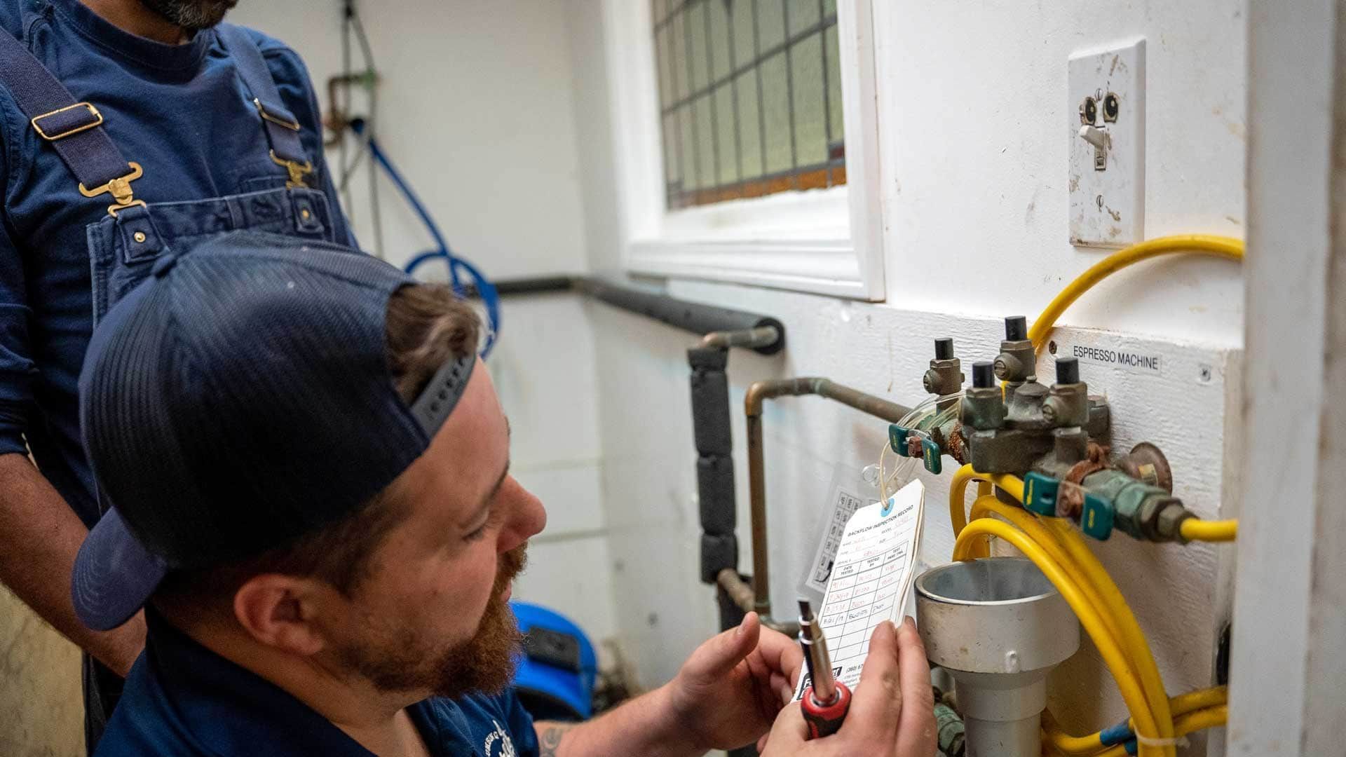 Two men working on plumbing in a utility room. One man examines a checklist.
