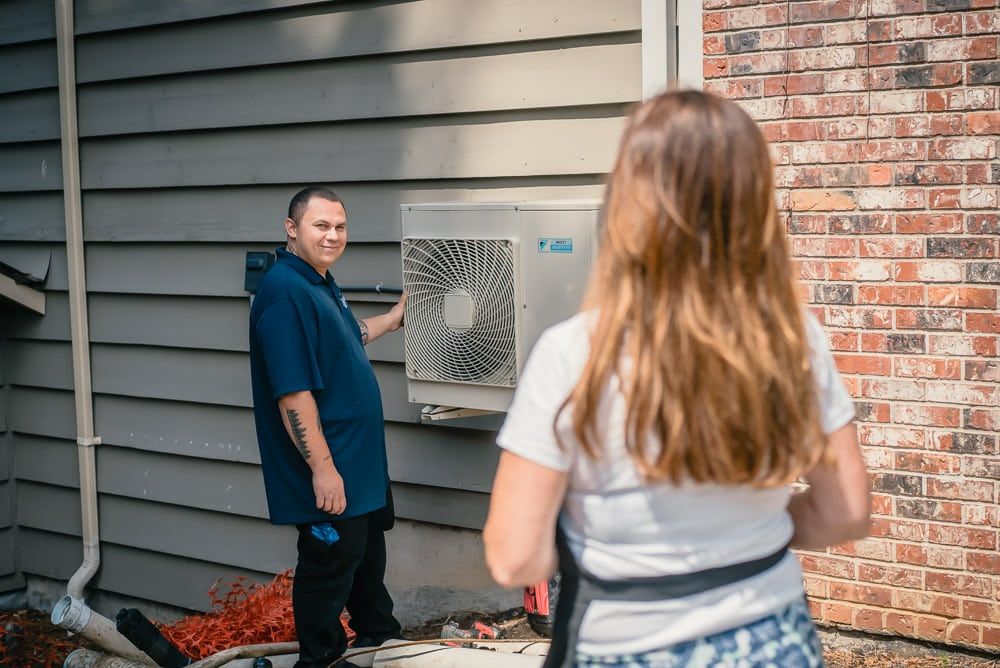 A woman is taking a picture of a man installing an air conditioner on the side of a house.