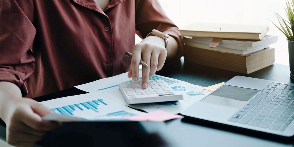 A woman is sitting at a desk using a calculator and a laptop.