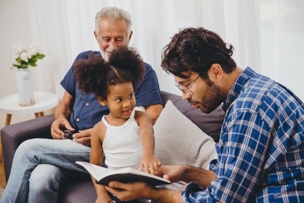 A man is reading a book to a little girl while two men sit on a couch.