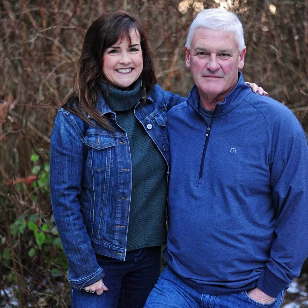 Woman in jean jacket and man in blue shirt pose outdoors.