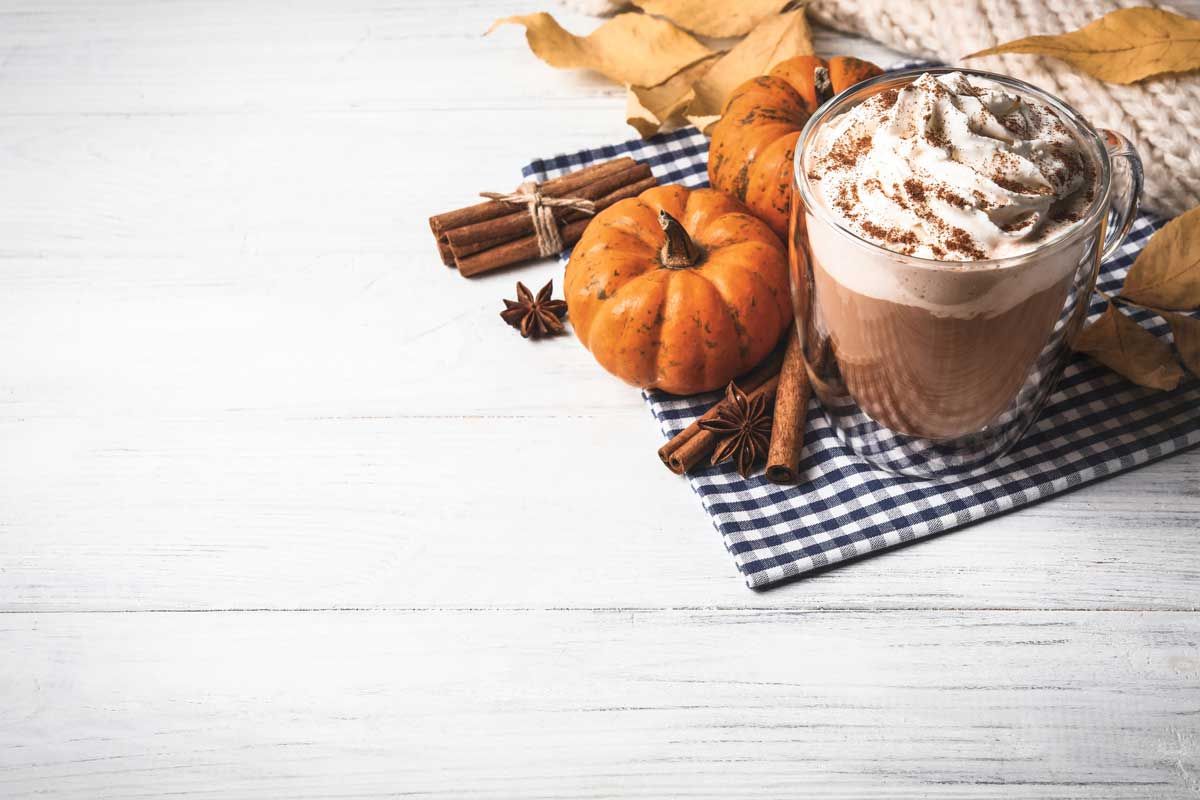 Pumpkin spice latte with pumpkins, cinnamon sticks, and autumn decor on a white wooden table.