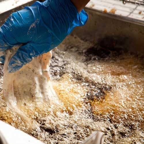 A gloved hand dips a breaded item into bubbling hot oil for frying in a metal container.