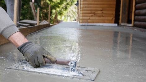 Person using a trowel to smooth wet concrete on a porch. Gray gloves, building in the background.