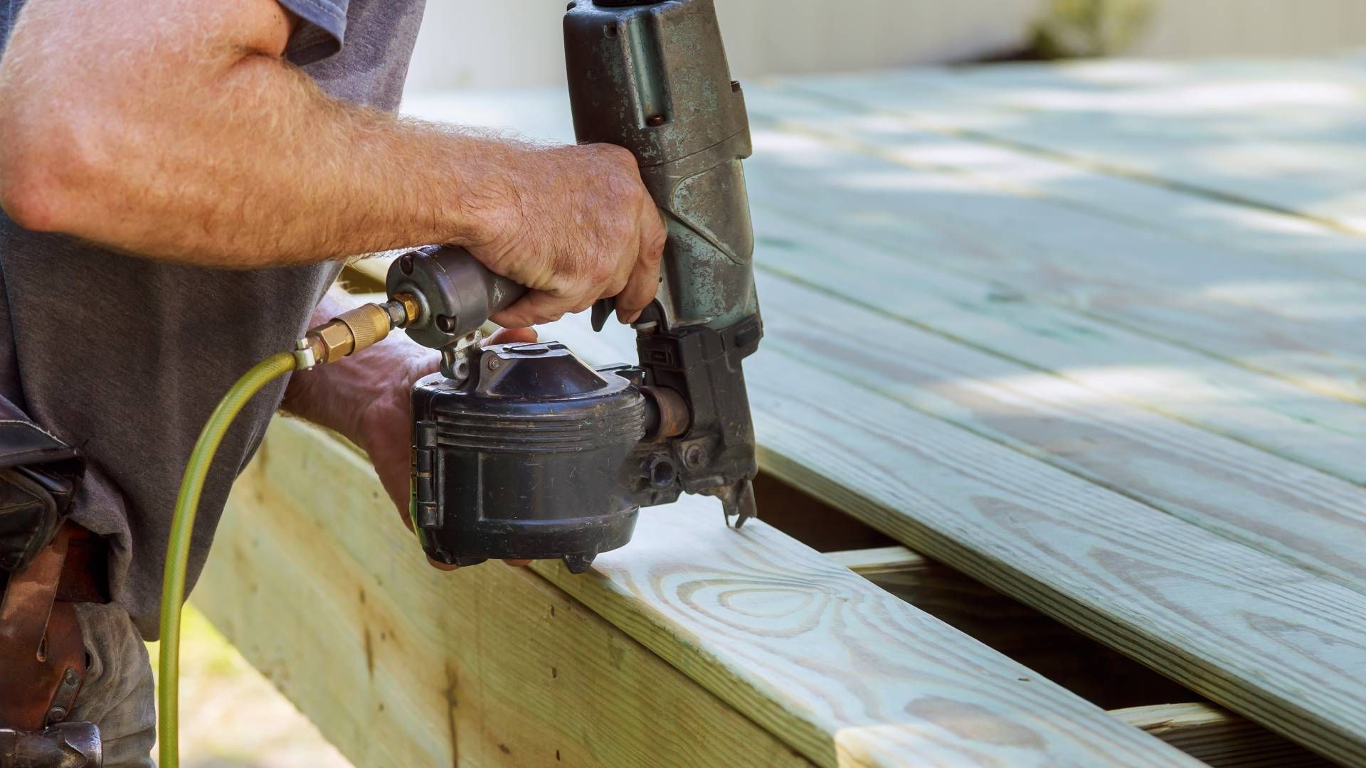 Person using a nail gun to attach wooden planks to a deck frame outdoors.