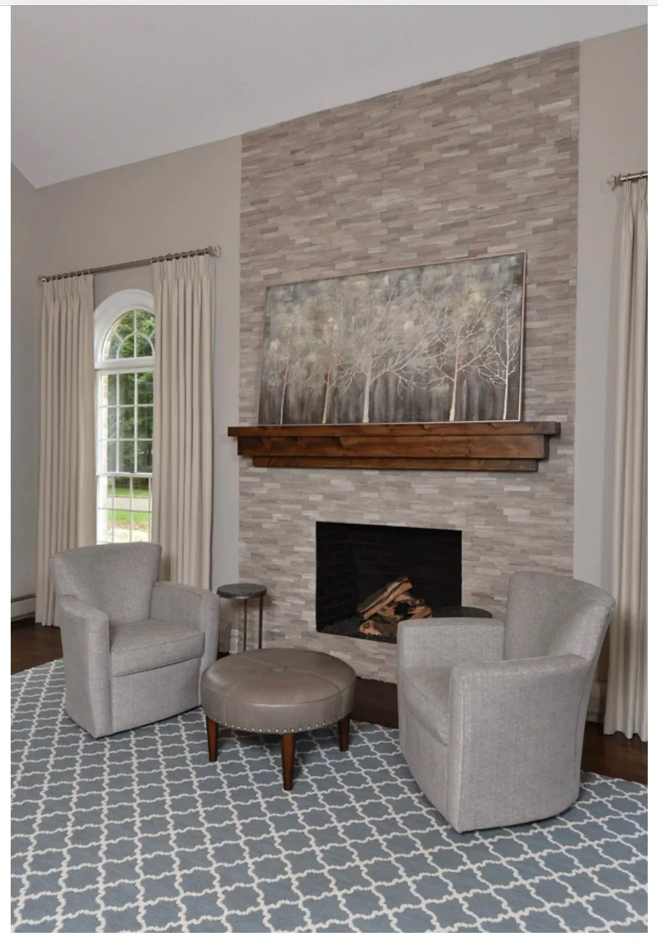 Living room with a fireplace, two gray armchairs, and an ottoman on a blue patterned rug.