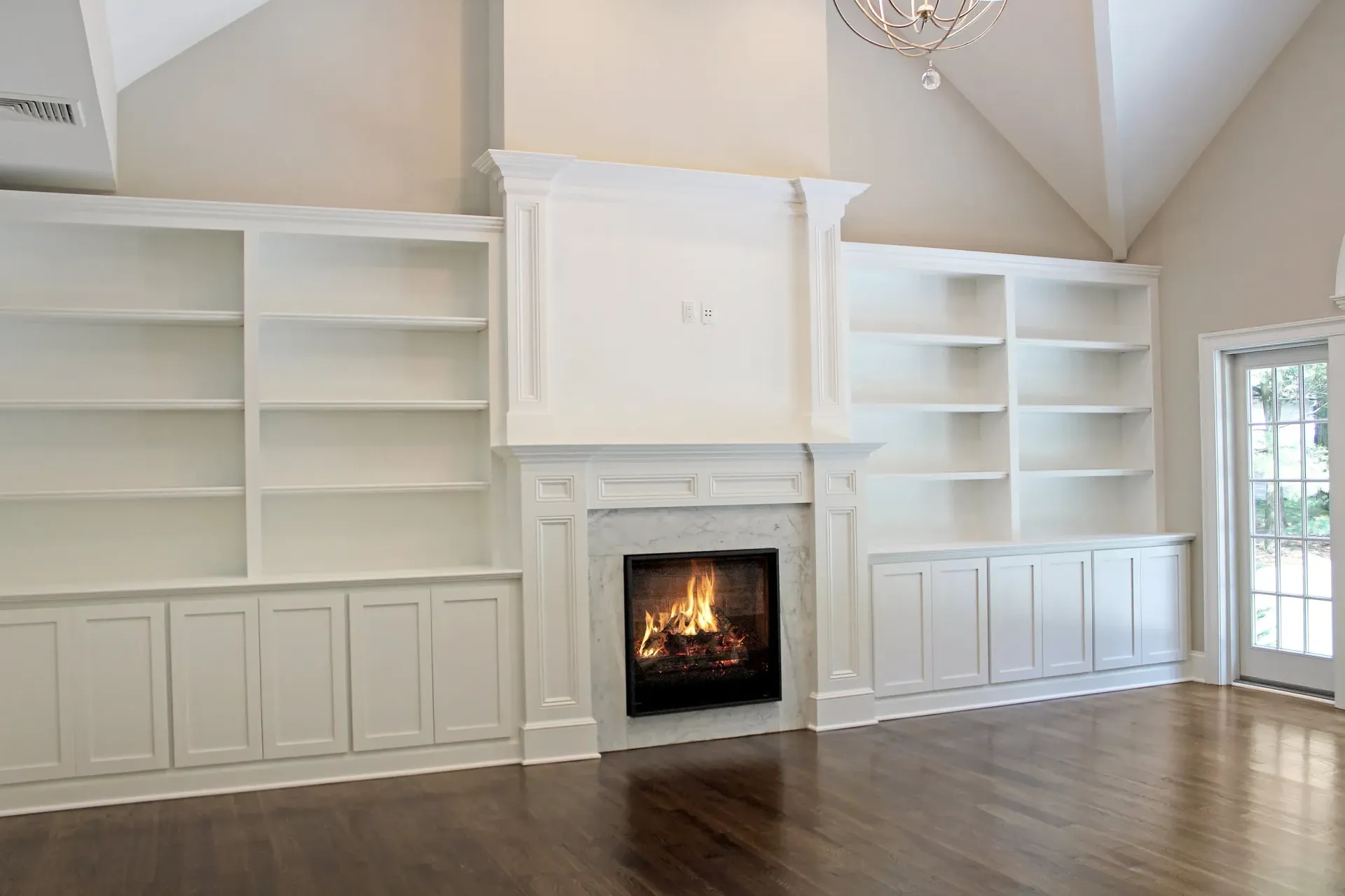 White built-in shelves and fireplace surround a fireplace with a burning fire in a room with dark wood floors.