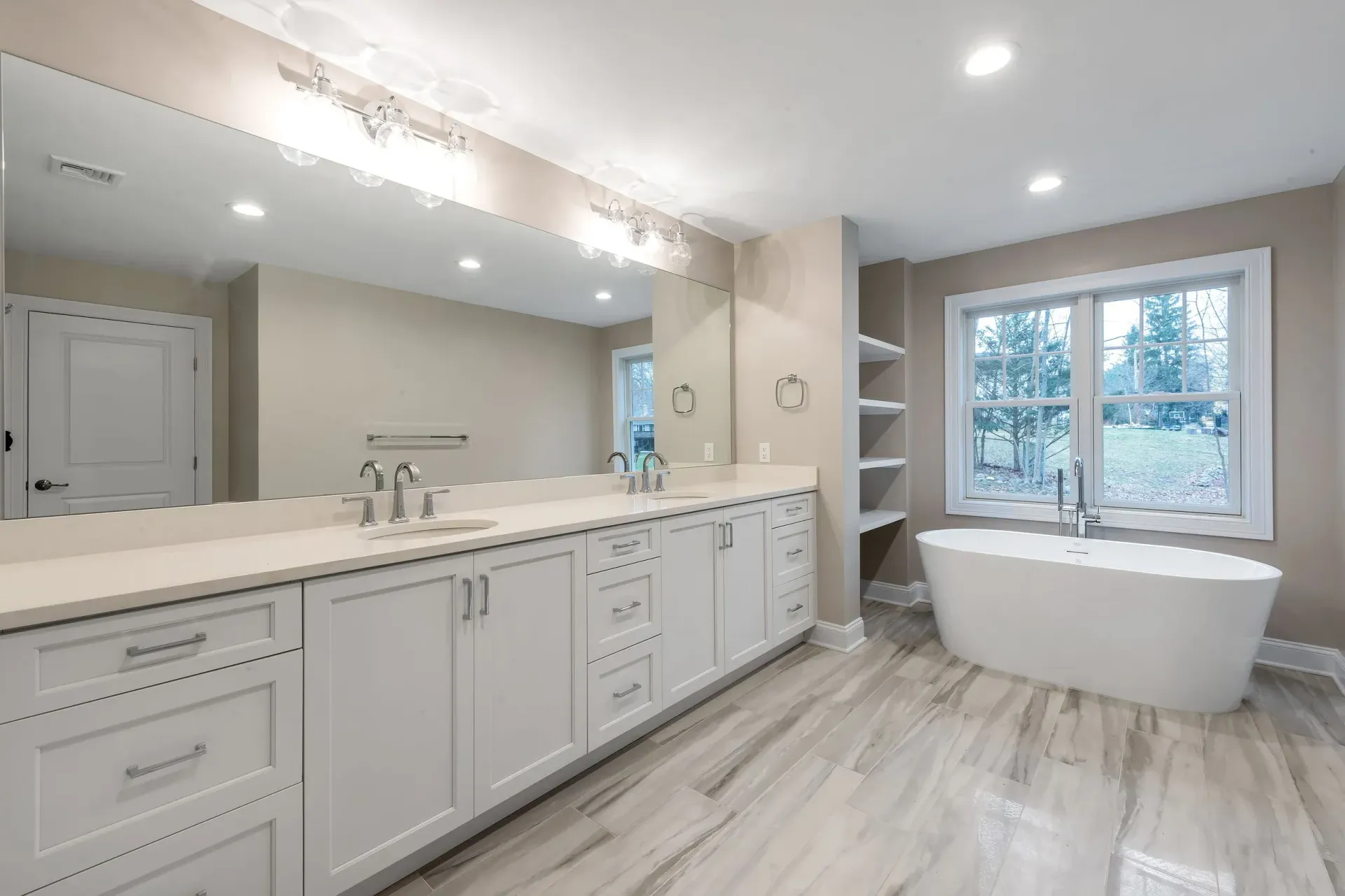 Spacious white bathroom with a long vanity, a standalone tub, and a window with outdoor views.