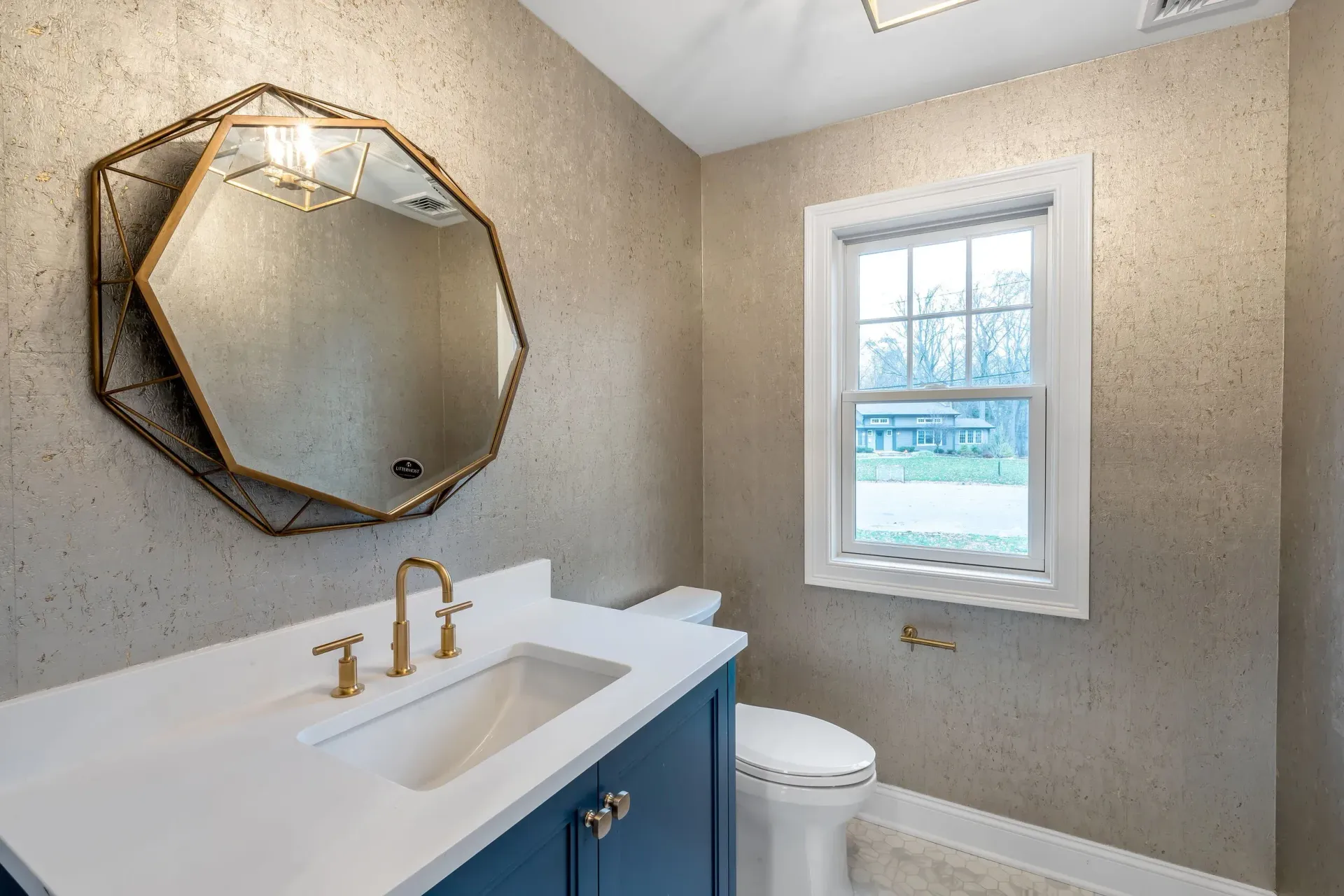 Bathroom with blue vanity, gold fixtures, patterned wallpaper, and an octagonal mirror.