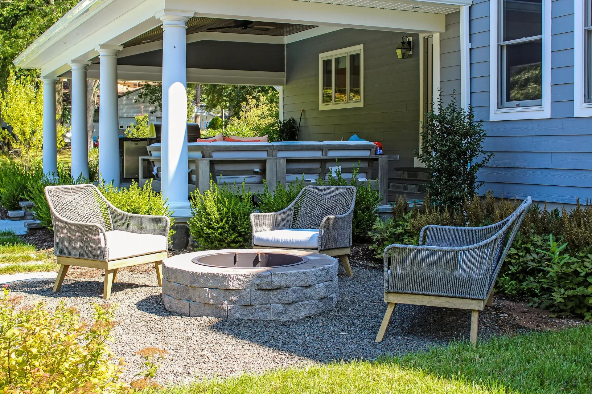 Outdoor seating area with fire pit and chairs in front of a blue house with a porch.
