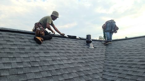 Two roofers installing shingles on a gray shingled roof under a cloudy sky.