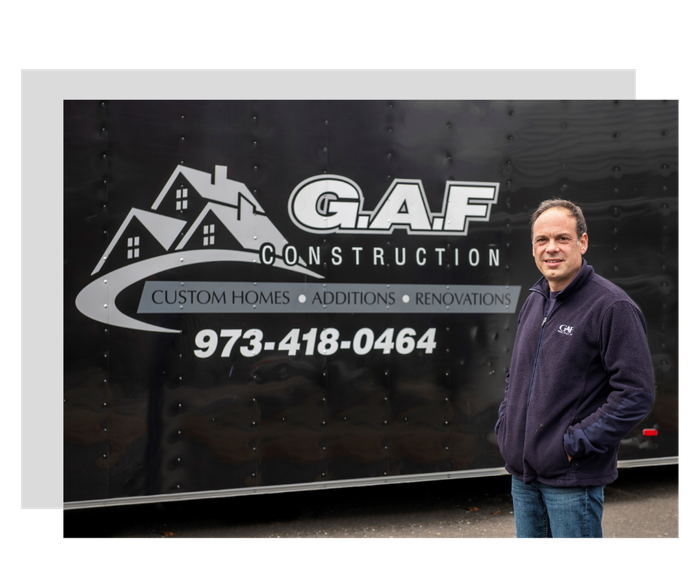 Man standing next to a black GAF Construction trailer. The trailer displays the company logo and contact information.