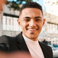 A young man in a suit and white turtleneck is smiling.