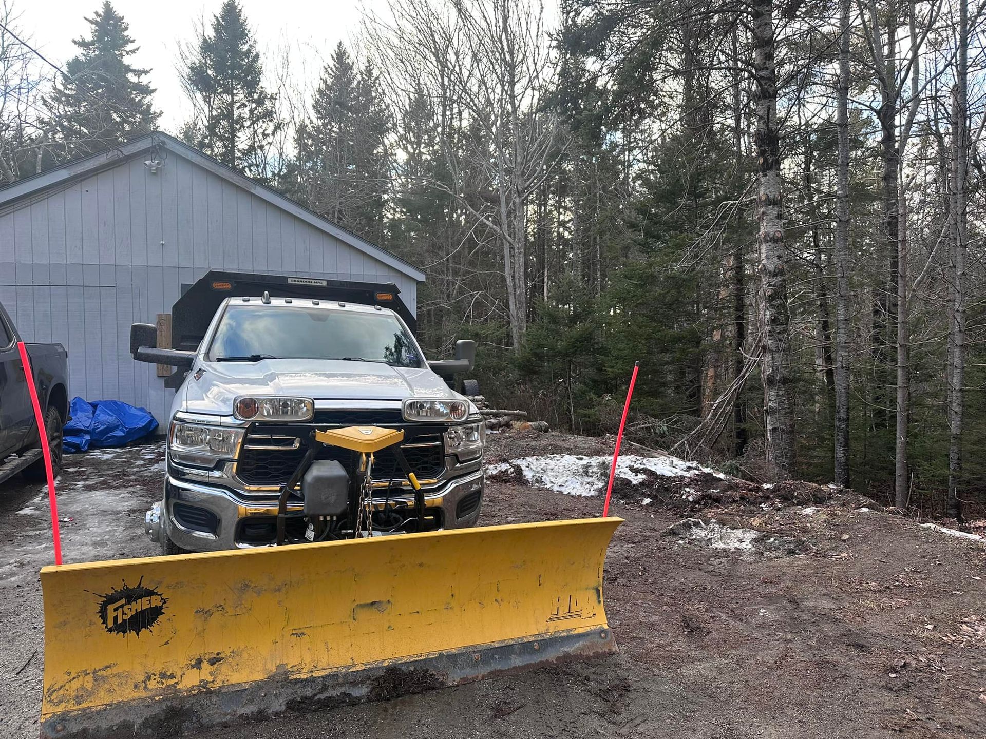 A truck with a yellow snowplow is parked in front of a light blue garage. Trees in the background.