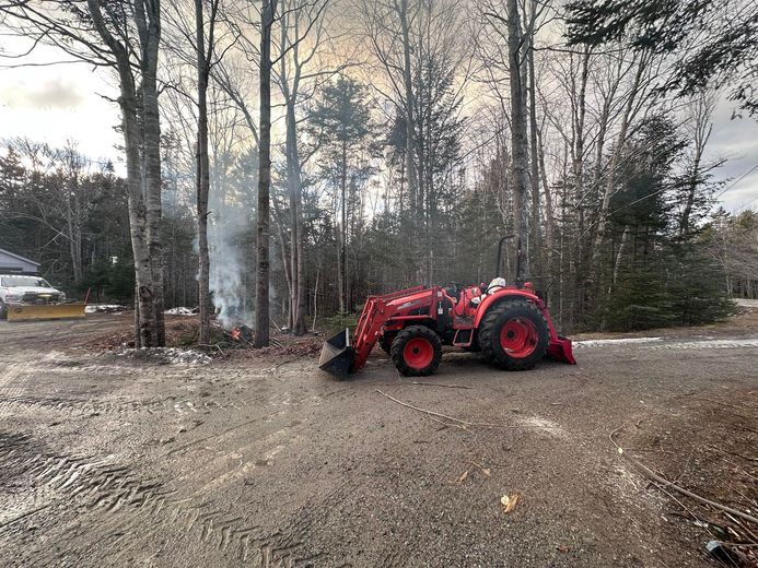 Red tractor with loader parked on gravel near a burning pile in a wooded area.