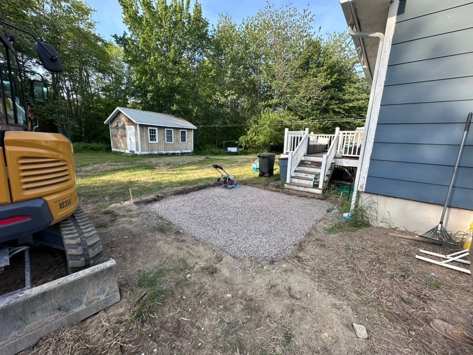 Gravel patio next to a blue house with wooden stairs; small shed in the background, excavator on the left.