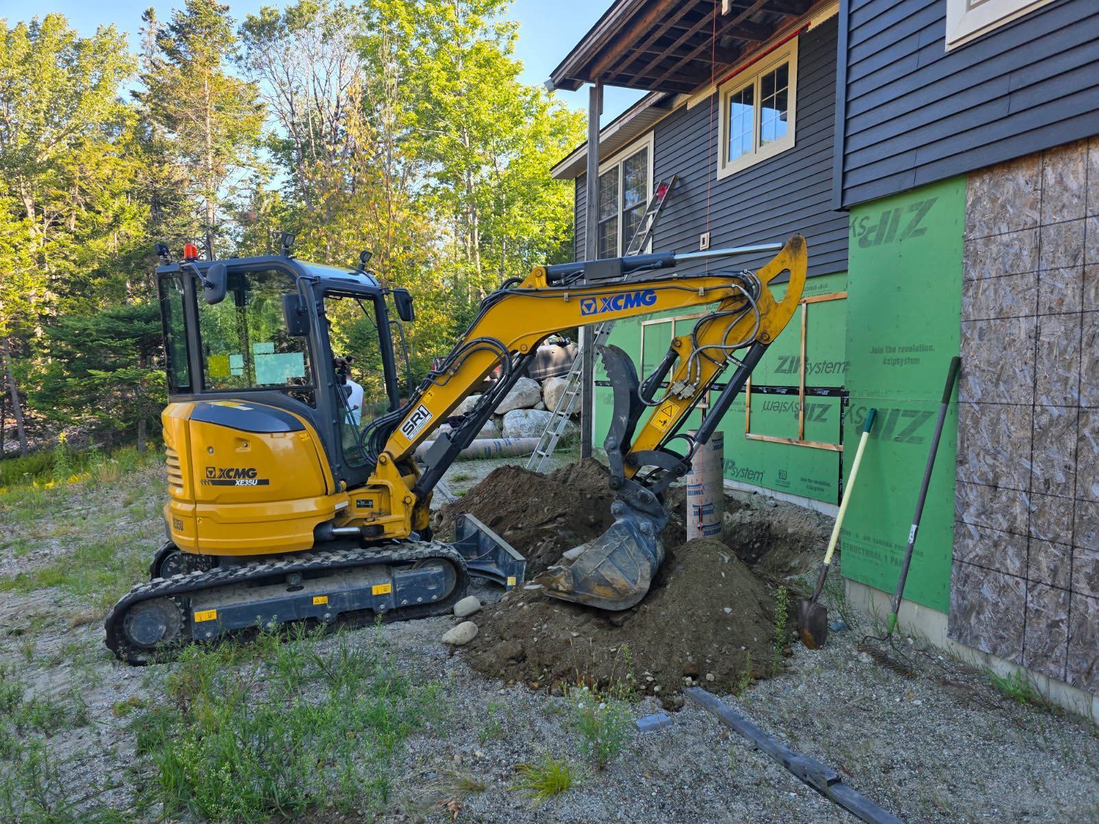 Yellow excavator digging near a house with green siding.