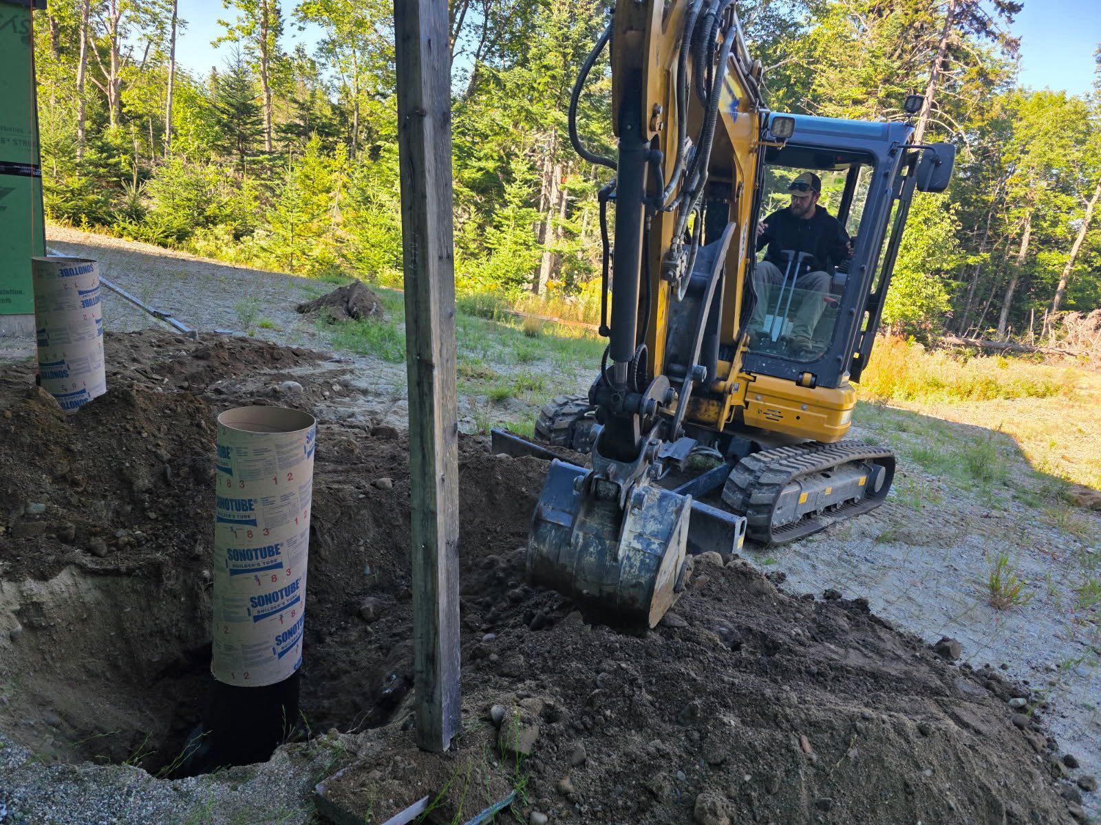 An excavator digs a hole next to a building; a man operates the machine in an outdoor setting.