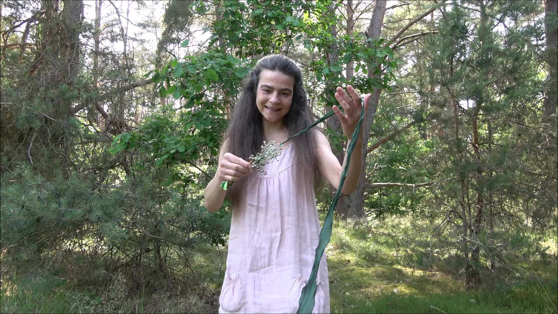 Eine Frau im rosanen Kleid steht im Wald und hält ein Zepter mit einem grünen Band