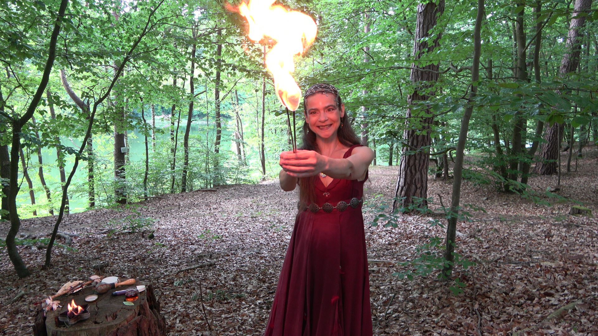 Eine Feuerfee im roten Kleid hält im Wald Feuerstäbe in der Hand