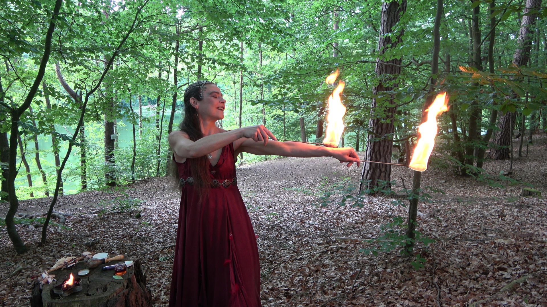 Eine Feuerfee im roten Kleid hält im Wald Feuerstäbe in der Hand
