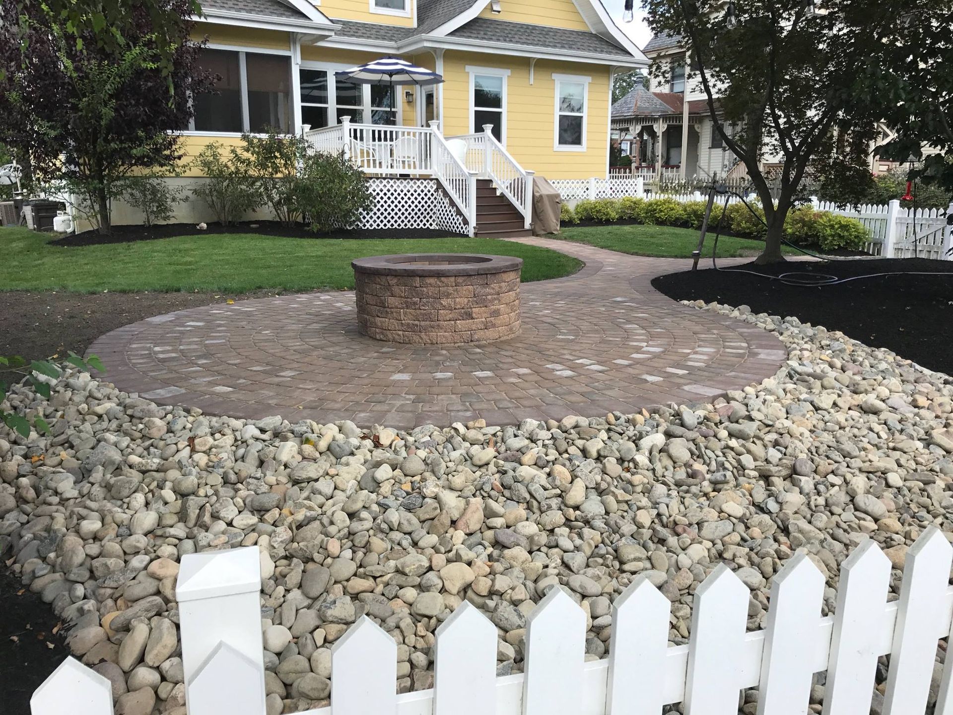 A white picket fence surrounds a fire pit in front of a yellow house