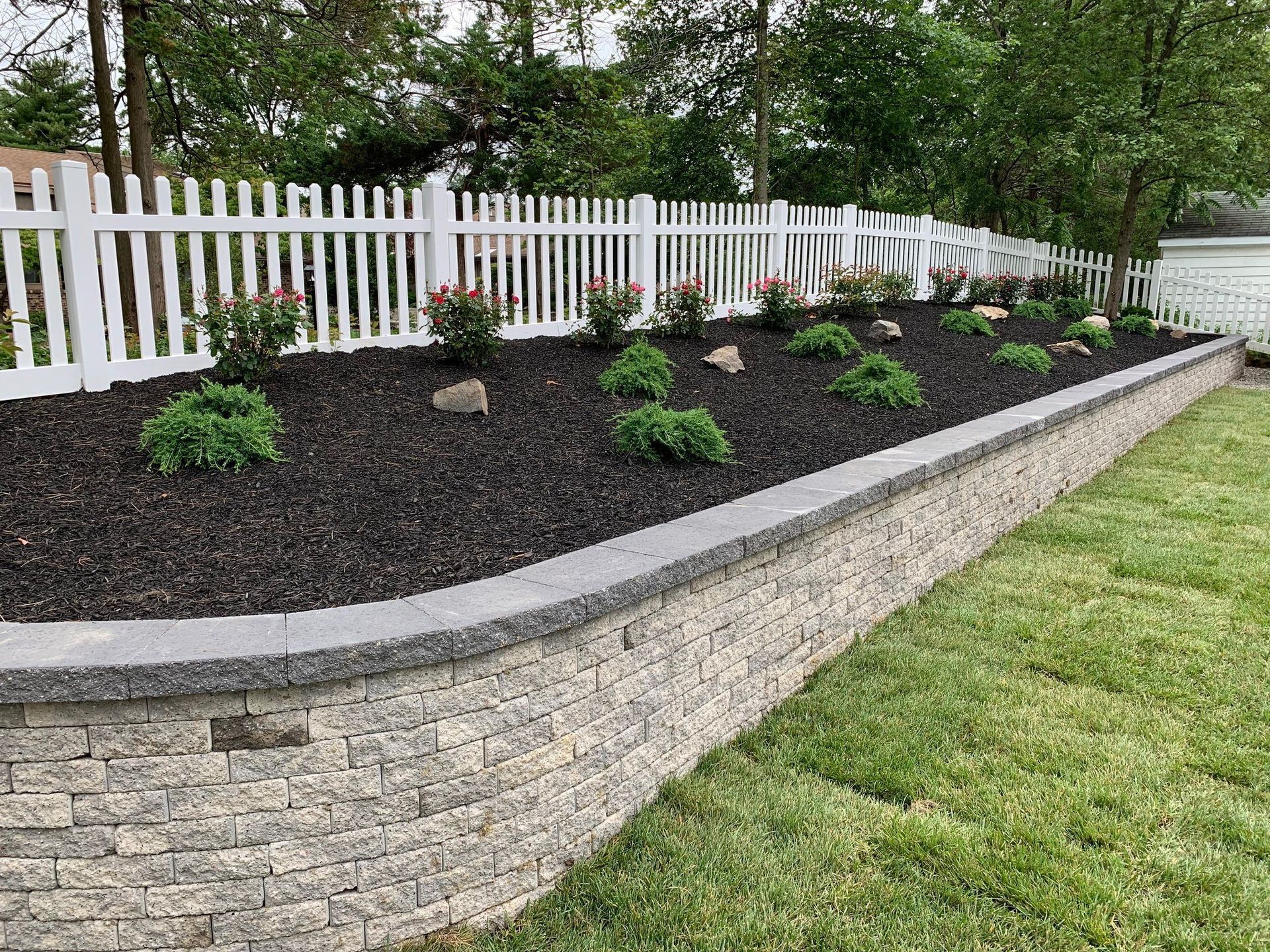 A white picket fence is surrounded by a brick wall and a garden.