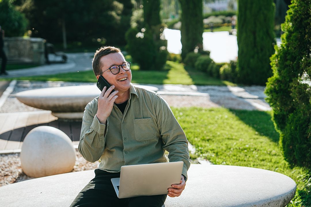 A man is sitting on a bench with a laptop and talking on a cell phone.