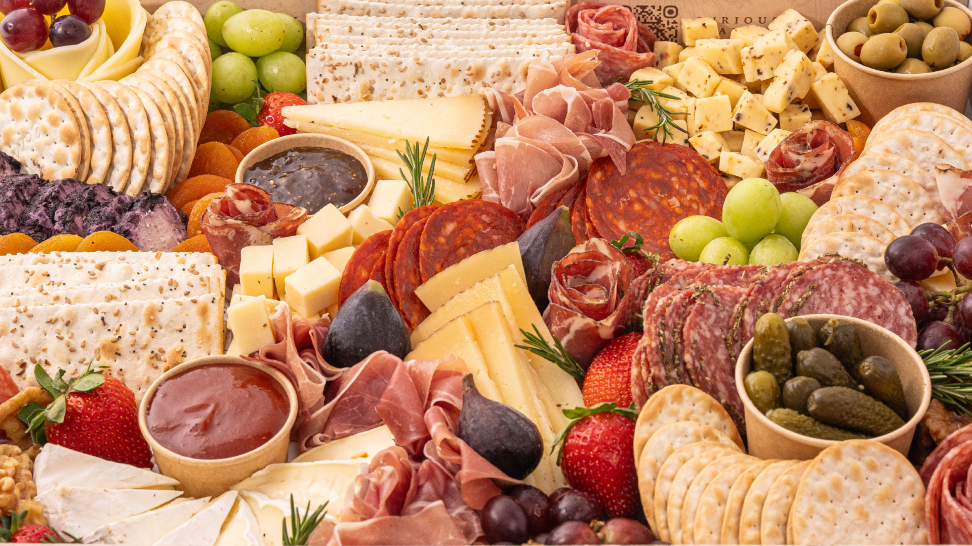 A variety of meats , cheeses , fruits and vegetables on a cutting board.