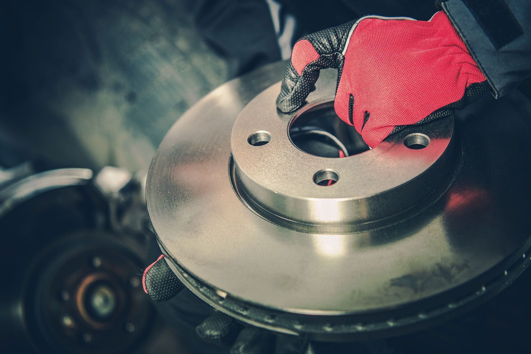 Mechanic holding a new brake rotor, wearing red gloves, in a garage.