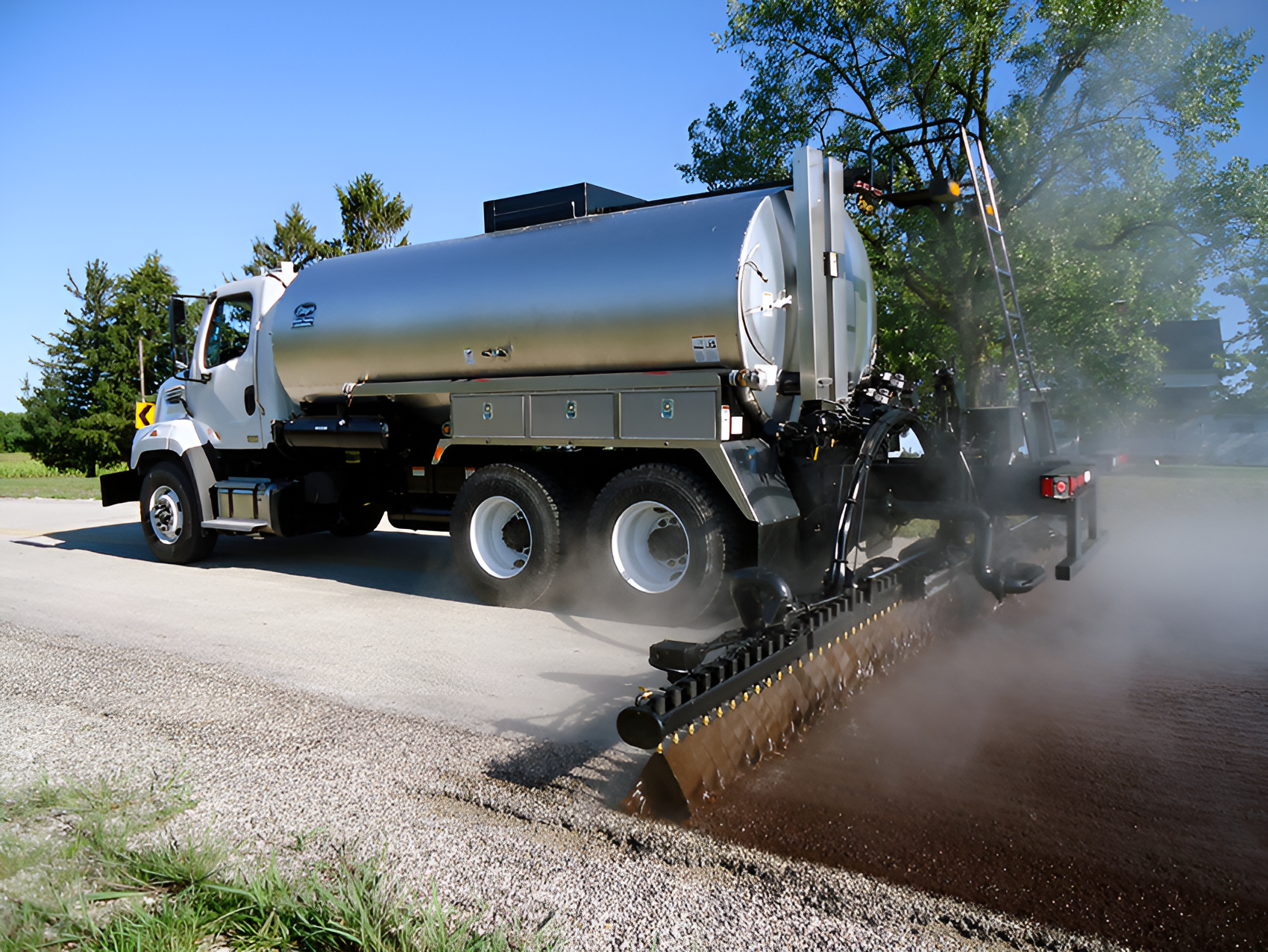 A silver truck is spraying asphalt on a gravel road