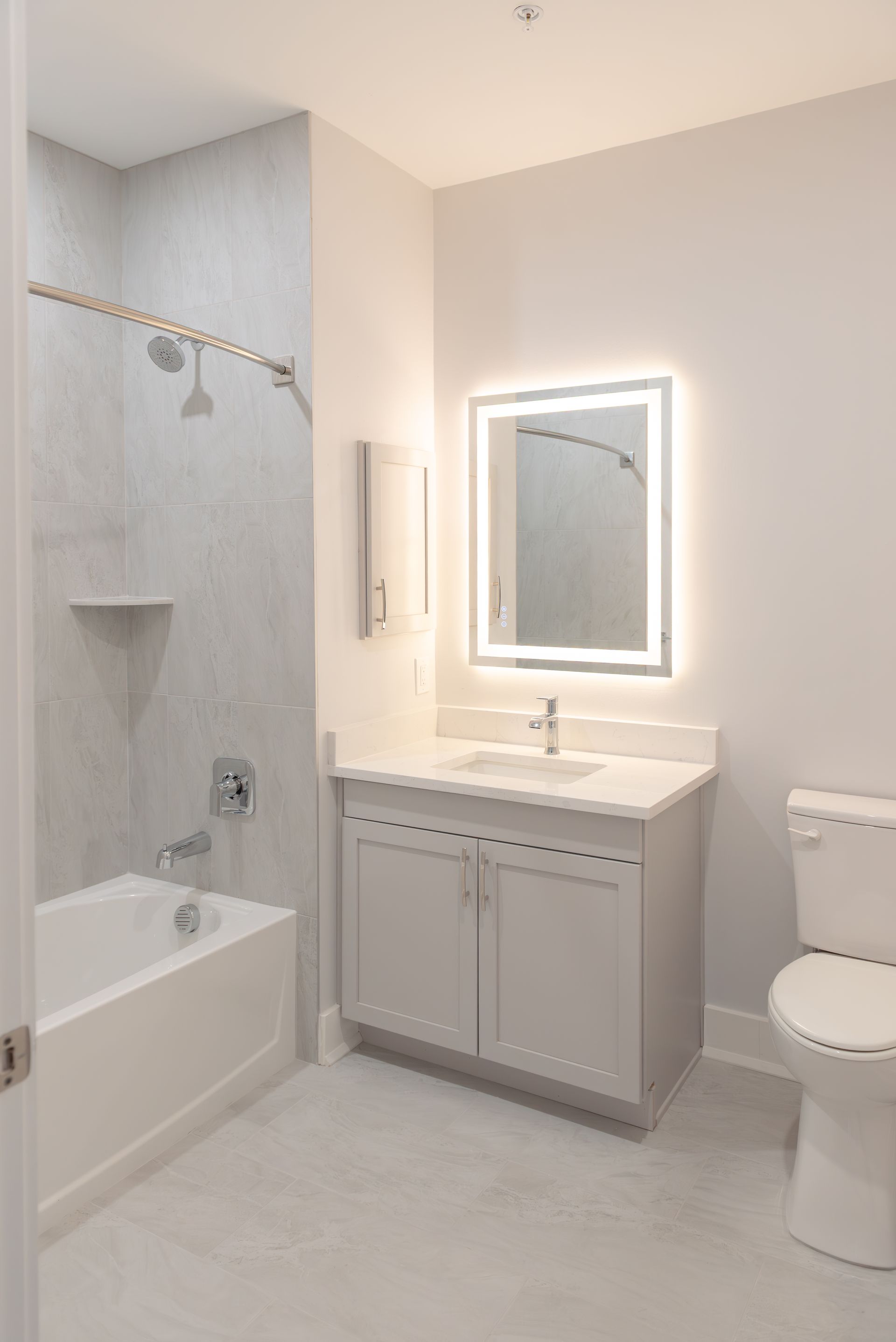 Bright bathroom with white fixtures, gray walls, and a lighted mirror.