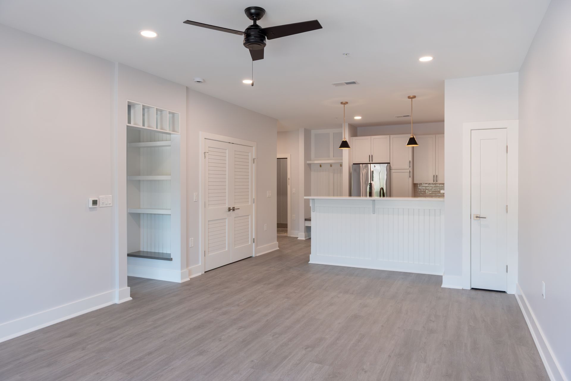 Empty, bright, modern living space with a kitchen. Light grey wood-look floors, white walls, cabinets, and trim. Ceiling fan.