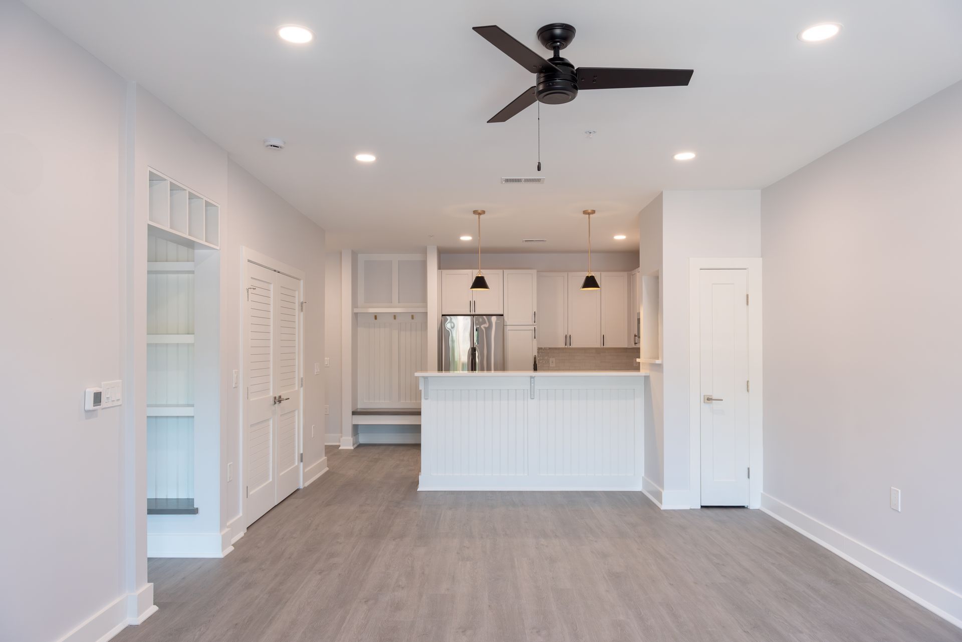 Spacious, bright interior with kitchen and living area. White walls, light grey flooring, and black ceiling fan.