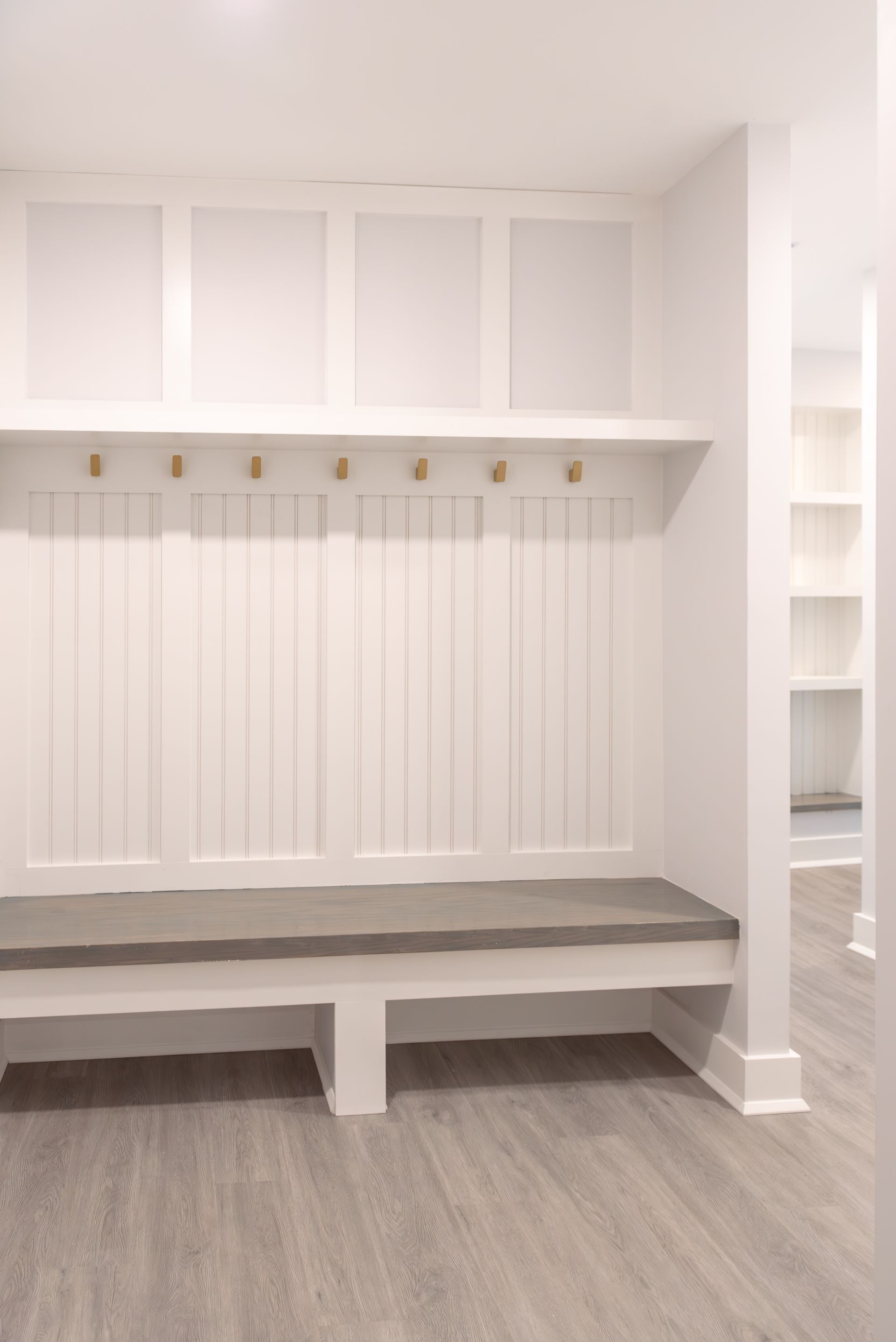White mudroom with bench, hooks, and built-in shelf. Light grey flooring and bright walls.